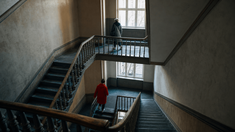 Zwei Personen unterschiedlichen Alters gehen auf verschiedenen Ebenen durch ein weitläufiges Berliner Treppenhaus.“